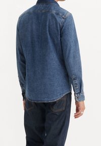 Camicia di jeans blu scuro, maniche lunghe con polsini abbottonati e orlo curvo. Presenta un colletto classico e un tessuto liscio e strutturato.