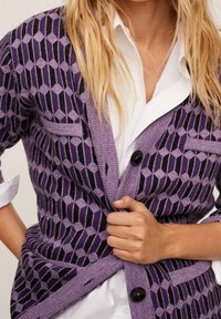 Žena v fialovém kardiganu s geometrickým vzorem přes bílou košili s knoflíky, zapíná prostřední knoflík kardiganu.