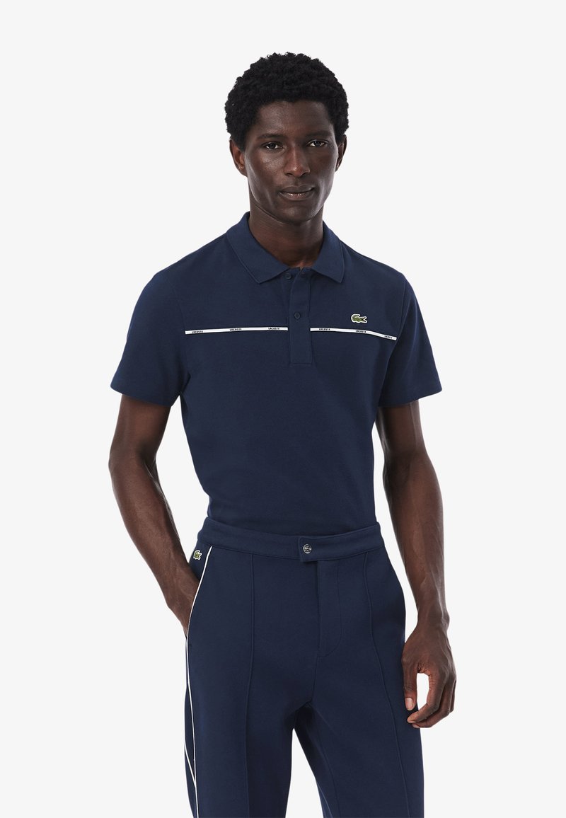 Mörkblå polotröja i bomull, med krage, korta ärmar och vita horisontella ränder över bröstet. Lacoste-logotyp på vänster sida.