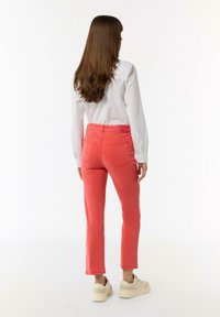 Coral pants with a slim fit and cropped hem, featuring two back pockets. Paired with a white long-sleeve shirt and beige footwear.