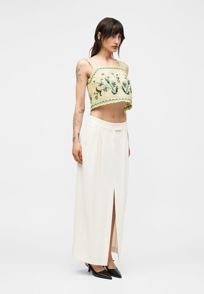 Γυναίκα με ανοιχτό κίτρινο κεντητό crop top, μακριά λευκή φούστα με σκίσιμο μπροστά και μαύρες μυτερές γόβες, στέκεται μπροστά σε απλό φόντο.