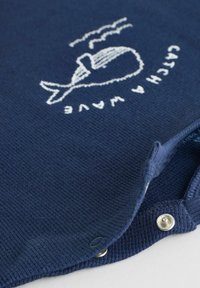 Pull en maille bleu marine avec une baleine brodée blanche et le texte « ATTRAPE UNE VAGUE », avec des boutons-pression au col.