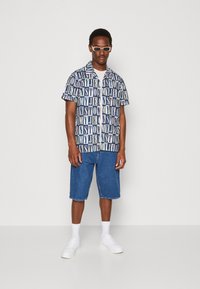 Camicia a maniche corte con bottoni, caratterizzata da un audace motivo di testo ripetuto blu e bianco, abbinata a pantaloni corti di denim e sneakers bianche.