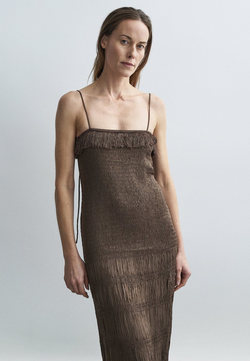 Barna, ujjatlan ruha, mely smockolt textúrával és rojtos részlettel rendelkezik a tetején. Hosszú, testhezálló fazon változó vízszintes redőkkel.