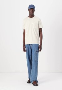 Cream cotton t-shirt with a chest pocket, paired with light blue denim cargo pants featuring side pockets, and a blue cap. Brown shoes.