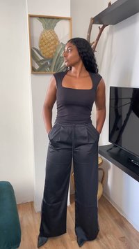 Pikkade lokkis juustega naine mustas toppis ja laia säärega pükstes seisab käed taskutes, vaadates kõrvale elutoas.