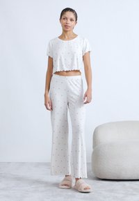 Λευκό σετ ρούχων για χαλάρωση που περιλαμβάνει ένα cropped τοπ με κοντά μανίκια και κυματιστό τελείωμα, σε συνδυασμό με αέρινο παντελόνι διακοσμημένο με μικρές κόκκινες καρδιές.