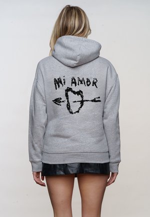Femme portant un sweat à capuche gris avec un cœur graphique noir traversé par une flèche et le texte « Mi Amor » au dos, associée à une jupe noire.