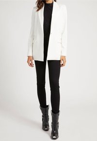 Blazer blanc à coupe ajustée, avec des revers en encoche et deux poches avant, assorti à des leggings noirs et des bottines noires épaisses.