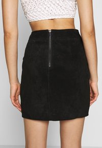 Mini-jupe en daim noire avec une fermeture éclair au dos, présentant une texture lisse et un design simple. Aucun motif visible ni accent supplémentaire.