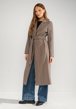 Femme portant un long manteau marron à carreaux ceinturé sur un jean bleu à jambes larges et des chaussures noires, debout devant un fond clair uni.