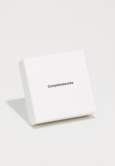 Λευκό τετράγωνο κουτί με κομψό σχεδιασμό και ματ υφή, που φέρει την επωνυμία "Completedworks" τυπωμένη με μαύρη γραμματοσειρά στο μπροστινό μέρος.