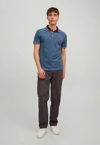 Polo blu con colletto rosso e dettagli rossi, abbinata a pantaloni grigio scuro. Il modello è in piedi con le mani lungo i fianchi, indossando scarpe da ginnastica bianche.