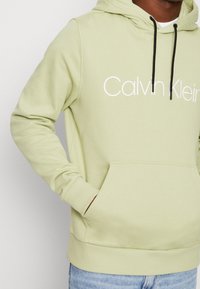 Ljusgrön hoodie i mjukt material, med ficka framtill, dragsko på huvan och vit "Calvin Klein"-logotyp på bröstet.