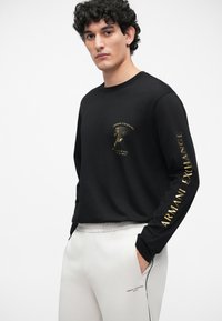 Melnas garām piedurknēm T-krekls ar zelta logotipu un teksta akcentiem. Apvienots ar gaiši pelēkiem treniņbiksēm ar mazu logotipu. Vienkārša dizaina, brīva piegriezuma.