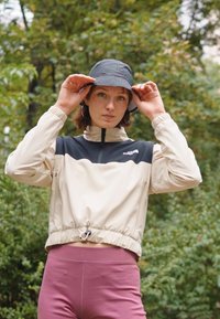 Rompevientos beige y negro con cuello alto, cintura elástica y cremallera. Combinado con leggings rosas, complementado con un sombrero negro tipo bucket.
