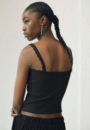 Débardeur noir à côtes avec des fines bretelles festonnées, design ajusté et texture lisse, porté avec un pantalon noir à cordon de serrage.