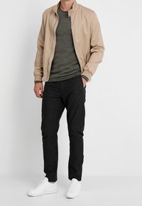 Blouson aviateur beige, chemise verte foncée texturée, pantalon noir fuselé et baskets blanches. Design simple, coupe confortable.