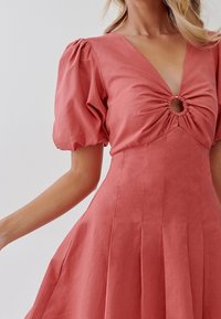 Šaty v rustové barvě s nabíranými rukávy a hlubokým výstřihem do V. Jsou vybaveny nabíraným středem s kulatým výřezem. Sukně je rozšířená a plisovaná.