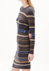 Femme portant une robe ajustée en tricot côtelé avec des rayures horizontales grises, bleues, beiges et brunes, se tenant devant un arrière-plan blanc.