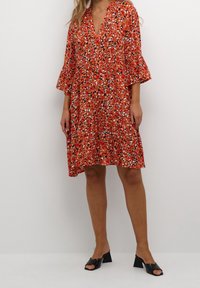 Červené šaty s květinovým vzorem, výstřihem do V, tříčtvrtečními zvonovými rukávy a volným střihem. Opuštěno s černými podpatkovými pantofli.