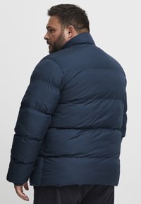 Navy blue padded jacket with a high collar and a horizontal quilted pattern. Features a relaxed fit and elastic cuffs. Soft, smooth texture.