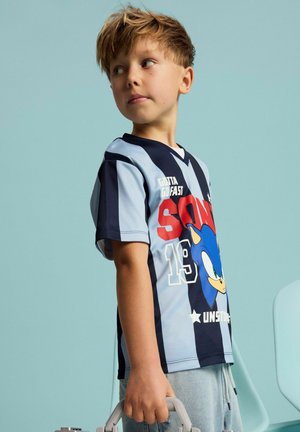 Nuori poika, jolla on päällään sinimustaraidallinen Sonic the Hedgehog -paita, katsoo sivulle ja pitää harmaata kahvaa vaaleansinistä taustaa vasten.