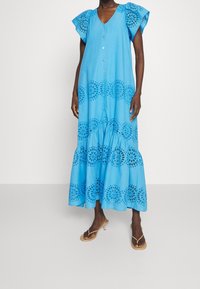 Robe bleue en coton, longueur genou, avec un col en V, des manches courtes volantées, des boutons sur le devant, et des motifs circulaires brodés à l'ourlet.