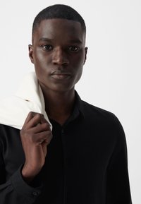 Camisa negra de botones con mangas largas, que presenta una textura suave. Tela color crema drapeada sobre el hombro, destacando el contraste.