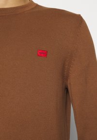 Pull en maille marron avec un col rond, arborant un petit patch rectangulaire rouge marqué "HUGO" sur la poitrine gauche.