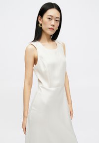 Αμάνικο κρεμ φόρεμα με απαλή σατέν υφή, που διαθέτει στρογγυλή λαιμόκοψη και προσαρμοσμένη εφαρμογή. Χρυσά σκουλαρίκια προσθέτουν λεπτομέρεια.