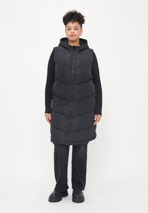 Schwarze Steppweste mit Kapuze, ausgestattet mit einem Reißverschluss an der Vorderseite, gestepptem Design und langer Länge. Getragen über einem schwarzen Langarmshirt und einer Hose.