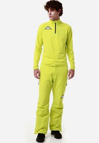 Completo sportivo di un giallo acceso composto da una maglia a maniche lunghe con zip a quarto e pantaloni coordinati, con un logo sul petto e sulla gamba.