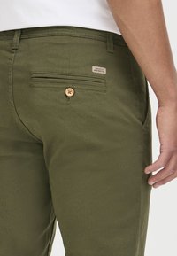 Gros plan d'une personne portant un pantalon vert olive avec une poche arrière boutonnée et une petite étiquette en tissu au-dessus de la poche.