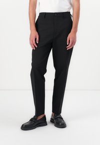 Čierne slim-fit nohavice z hladkej látky s gombíkovým zapínaním, bočnými vreckami a zoštíhleným strihom, doplnené čiernymi slip-on topánkami.