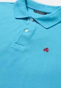 Tyrkysové polo tričko z texturované tkaniny má dvouknoflíkovou légu a červené vyšívané logo na hrudi. Klasický design límce.