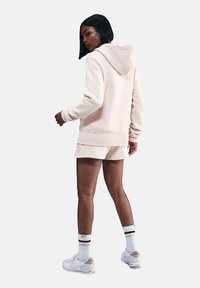 Felpa rosa chiaro con vestibilità morbida, abbinata a pantaloni corti coordinati. Presenta un cappuccio con coulisse e polsini a costine, indossata con scarpe da ginnastica bianche.