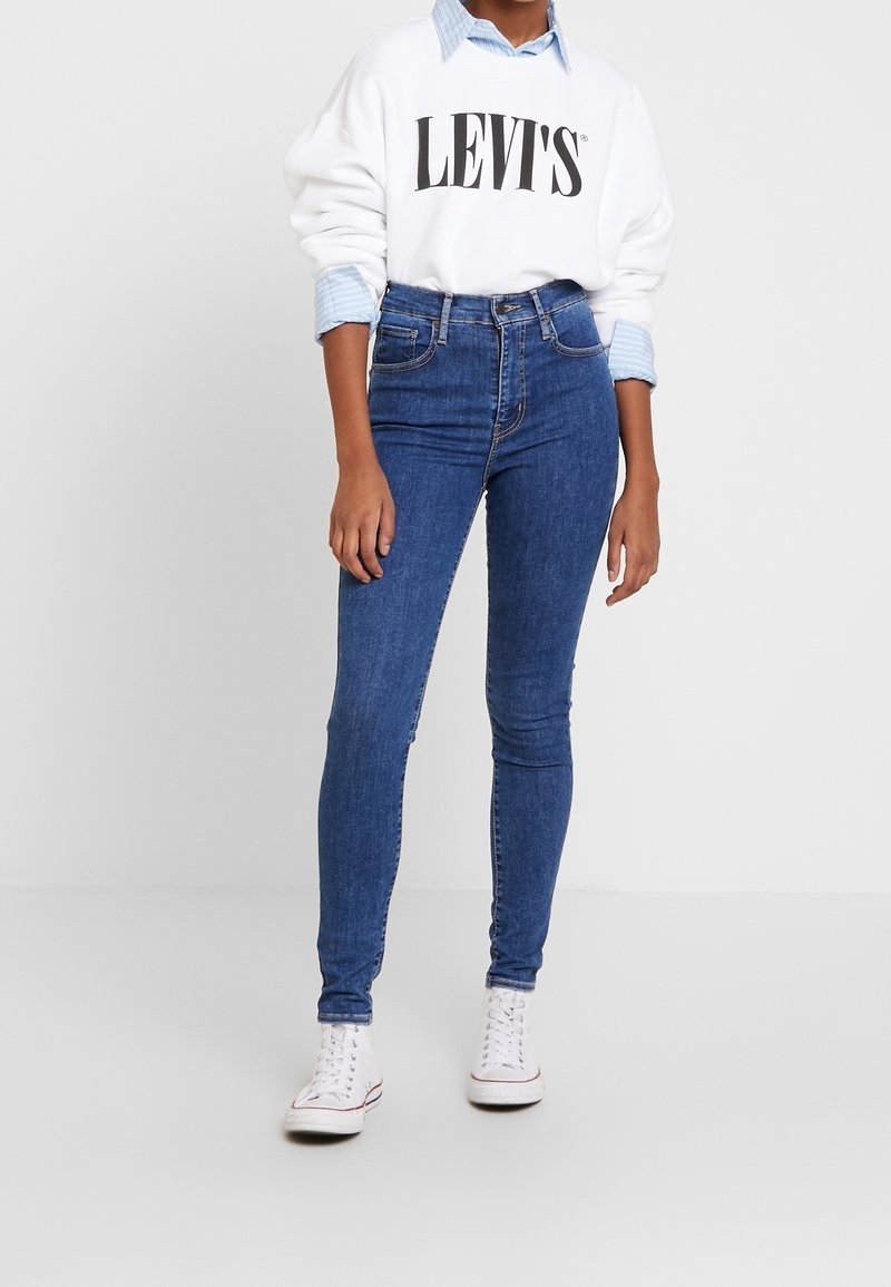 Personne portant un sweat-shirt blanc Levi's par-dessus une chemise bleue, un jean skinny bleu taille haute et des baskets blanches à lacets sur fond blanc.
