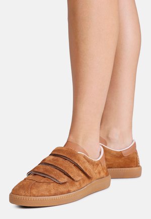 Καφέ πάνινα παπούτσια από σουέντ με τρεις λουρίδες βέλκρο, φορεμένα σε γυμνά πόδια σε απλό ανοιχτό χρωματικό φόντο.