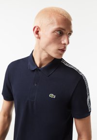 Marinblå pikétröja med två knappslå, korta ärmar och en grön broderad krokodil-logotyp. Har kontrasterande vita sidestreck.