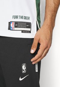 Maillot de basketball blanc avec l'inscription "FEAR THE DEER" en vert, logo Nike et logo NBA. Jogging noir avec logo NBA blanc et virgule Nike.