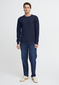 Navy blue knitted sweater with a crew neck and textured pattern, paired with dark denim jeans and white sneakers with gold accents.