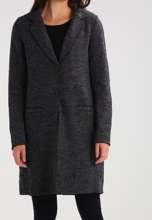 Vrouw draagt een donkergrijze wollen lange jas met knopen, reverskraag en voorzakken over zwarte kleding.