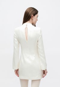 Witte, textuurrijke jurk met lange mouwen, een hoge halslijn en een open rug met een sleutelgat. Heeft een subtiele glans en een ritssluiting aan de achterkant.