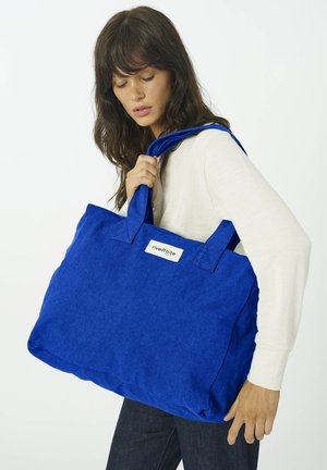 Femme aux longs cheveux foncés portant un haut blanc et un pantalon foncé, avec un grand sac fourre-tout bleu marqué "rivedroite" sur l'épaule.
