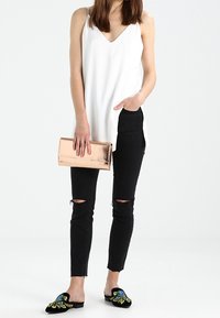 Mujer vistiendo una blusa sin mangas blanca, jeans ajustados negros desgastados, zapatos deslizables negros bordados y sosteniendo un clutch metálico color oro rosa.