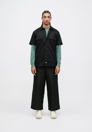 Chemise noire à manches courtes avec deux poches à l'avant, superposée à un haut teal à manches longues, associée à un pantalon noir large matelassé et des baskets claires.