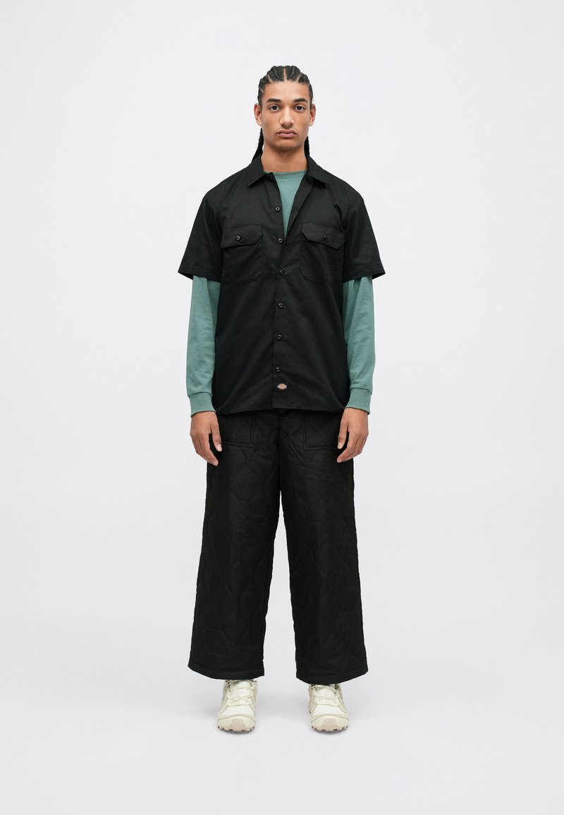 Černá košile s krátkým rukávem a dvěma předními kapsami, přepnutá přes dlouhý teplý top v barvě tmavé modro-zelené, v kombinaci s prošívanými širokými černými kalhotami a světlými teniskami.