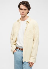 Ανοιχτό κίτρινο πουκάμισο με κουμπιά και διακριτική ριγέ υφή, φορεμένο πάνω από ένα λευκό T-shirt, συνδυασμένο με μπλε τζιν και καφέ ζώνη.