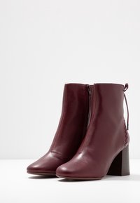 Botas de tobillo de cuero burdeos con puntera puntiaguda, cremallera lateral y detalles texturizados en el tacón. Tacones de bloque de madera; acabado suave.